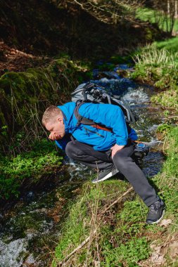 Yürüyüşçü dağda dere suyu içiyor. Yetişkin bir adam dağ deresinden su içiyor. Günlük giysiler ve güneş gözlüğü takan turist avuçlarından bir yudum nehir suyu içiyor.