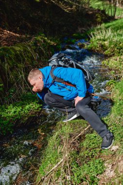 Yürüyüşçü dağda dere suyu içiyor. Yetişkin bir adam dağ deresinden su içiyor. Günlük giysiler ve güneş gözlüğü takan turist avuçlarından bir yudum nehir suyu içiyor.