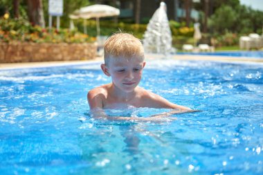 Küçük bir çocuk, sekiz yaşında, havuzda su sıçratıyor, eğlenceli aktiviteler yapıyor. Çocuk havuzda yüzmekten mutlu. Havuzda etkinlikler, yüzen ve suda oynayan çocuklar, mutluluk ve...