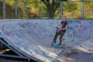 Parkta scooter kullanan bir çocuk. Çocuklar paten kaymayı öğreniyor. Küçük çocuk güneşli yaz gününde paten kayıyor. Güvenli yerleşim yerlerindeki çocuklar için açık hava aktivitesi. Anaokulu çocuğu için aktif spor. Çocuk