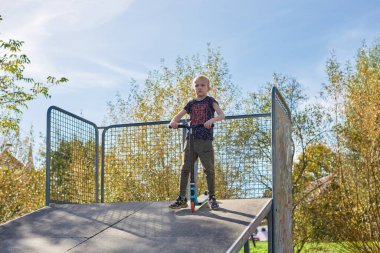 Scooter süren çocuk bir numara yapıyor ve bulutlu bahar gününde kaykay parkında eğleniyor. Genç adam parktaki scooter 'da numara yapıyor. Güneşli havada tüm vücuduyla scooter 'a bindi.
