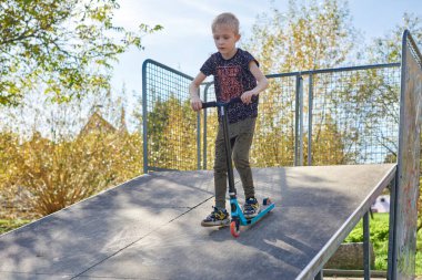 Scooter süren çocuk bir numara yapıyor ve bulutlu bahar gününde kaykay parkında eğleniyor. Genç adam parktaki scooter 'da numara yapıyor. Güneşli havada tüm vücuduyla scooter 'a bindi.