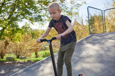 Parkta scooter kullanan bir çocuk. Çocuklar paten kaymayı öğreniyor. Küçük çocuk güneşli yaz gününde paten kayıyor. Güvenli yerleşim yerlerindeki çocuklar için açık hava aktivitesi. Anaokulu çocuğu için aktif spor. Çocuk