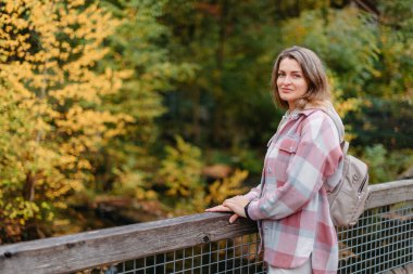 Sonbaharda gündelik giyinmiş genç bir kadının portresi, sonbahar parkının ve nehrin arka planında köprü üzerinde duruyor. Golden Fall 'da Park' ta yürüyen güzel bir kadın. Uzayı kopyala Gülümseyen kız