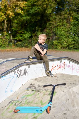 Parkta scooter kullanan bir çocuk. Çocuklar paten kaymayı öğreniyor. Küçük çocuk güneşli yaz gününde paten kayıyor. Güvenli yerleşim yerlerindeki çocuklar için açık hava aktivitesi. Anaokulu çocuğu için aktif spor. Çocuk