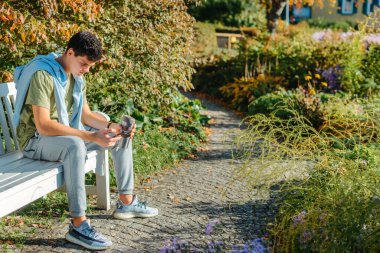 Parktaki bankta oturan bir genç termo bardaktan kahve içer ve telefona bakar. Yakışıklı, neşeli bir adamın portresi. Temiz hava almak için bankta oturuyor. Medya programında içki içiyor.