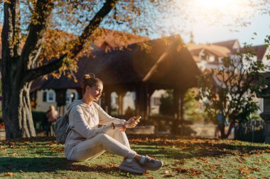 Sonbaharda parkta akıllı telefonlu genç ve şık bir kız gülümseyerek oturuyor. Sonbaharda parkta mesajlaşan modaya uygun bir genç kadın. Yenilenmiş, canlı renkler. Sıradan giyinen güzel sarışın genç kız.