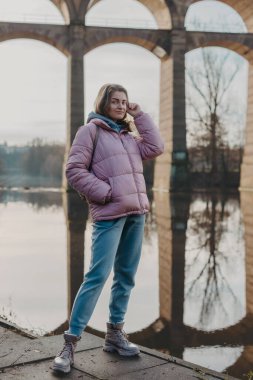 Sonbahar güneşli bir günde kıyıda durmuş nehri seyreden genç bir kadın. Sokak manzarası, metin için kopyalama alanı, seyahat fotoğrafı. Sonbaharda nehrin kıyısında sıcak giysiler içinde mutlu bir turist kadın.