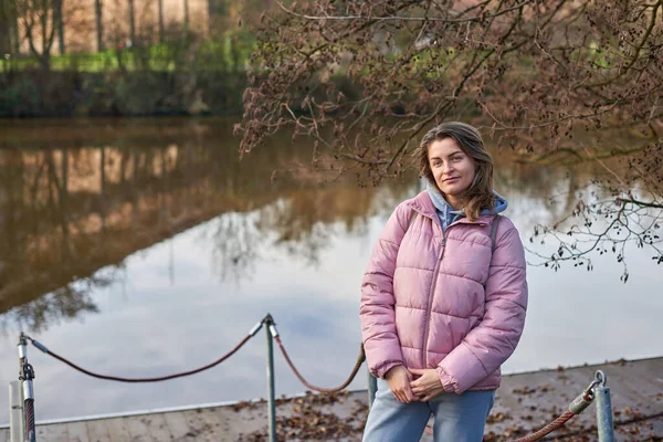 Sonbahar güneşli bir günde kıyıda durmuş nehri seyreden genç bir kadın. Sokak manzarası, metin için kopyalama alanı, seyahat fotoğrafı. Sonbaharda nehrin kıyısında sıcak giysiler içinde mutlu bir turist kadın.