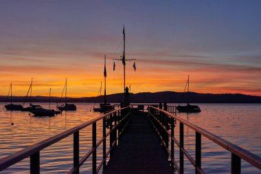 Bodensee Gölü Panorama. Akşam, alacakaranlık, batan güneş, manzara, sakin sular, rıhtımda tekneler ve yatlar, suda yansıyan bulutlar, alacakaranlıkta nehir kenarı.