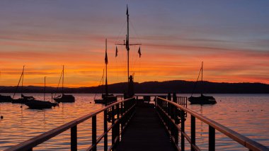 Bodensee Gölü Gündoğumu Panoraması. Sükunet Suları Üzerine Sabah Güneşi. Alman Bodensee Gölü üzerindeki büyüleyici şafağa tanık olun. Bir tekne iskelesinden yakalandılar. İlk zamanların huzurlu güzelliğini kucakla.