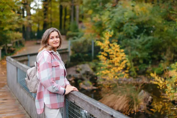 Portrait of cute young woman in casual wear in autumn, standing on bridge against background of an autumn Park and river. Pretty female walking in Park in golden fall. Copy space. smiling girl in the
