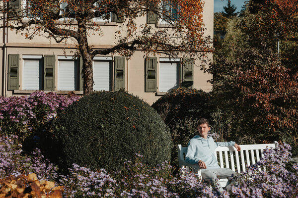 a teenager sits on a bench in the park drinks coffee from a thermo mug and looks into a phone. Portrait of handsome cheerful guy sitting on bench fresh air using device browsing media smm drinking