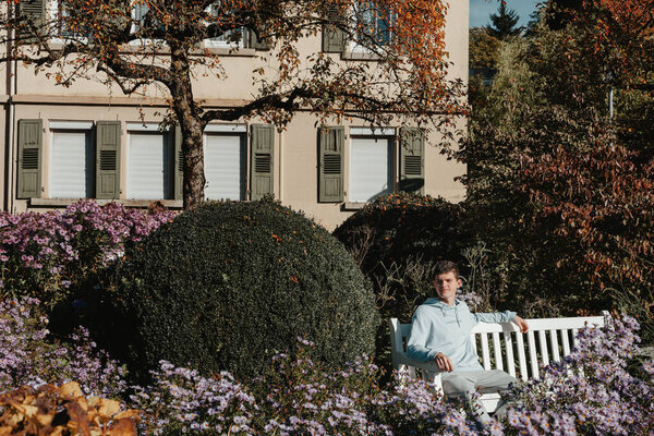 a teenager sits on a bench in the park drinks coffee from a thermo mug and looks into a phone. Portrait of handsome cheerful guy sitting on bench fresh air using device browsing media smm drinking