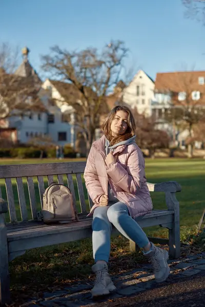 stock image Experience the festive spirit of winter with this delightful image capturing a beautiful girl in a pink winter jacket sitting on a bench in a park, set against the backdrop of the historic town of