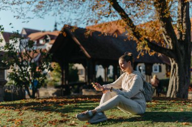 Sonbaharda parkta akıllı telefonlu genç ve şık bir kız gülümseyerek oturuyor. Sonbaharda parkta mesajlaşan modaya uygun bir genç kadın. Yenilenmiş, canlı renkler. Sıradan giyinen güzel sarışın genç kız.