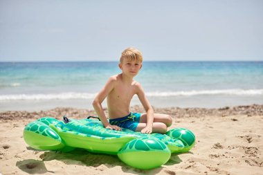 Tasasız Yaz Macerası: Genç Çocuk Şişme Yeşil Kaplumbağa Şamandırasında Dingin Okyanus Arkaplanlı Sunny Sandy Sahilinde Dinleniyor