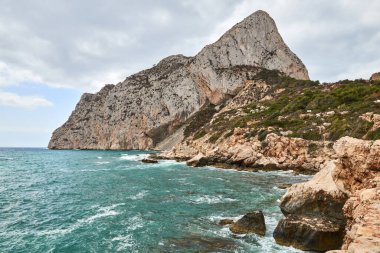 Calpe İspanya 'daki parlak güneşli gökyüzü altında kayalık uçurumlar ve turkuaz okyanuslarla sakin kıyı manzarası turizm için mükemmel.