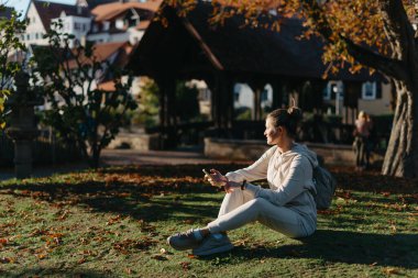 Sonbaharda parkta akıllı telefonlu genç ve şık bir kız gülümseyerek oturuyor. Sonbaharda parkta mesajlaşan modaya uygun bir genç kadın. Yenilenmiş, canlı renkler. Sıradan giyinen güzel sarışın genç kız.