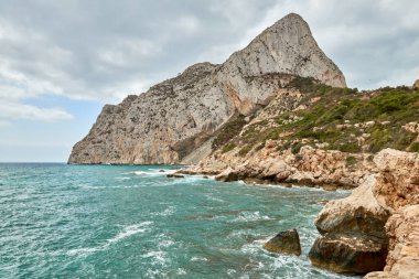 Gök mavisi sularıyla kıyı cenneti güneşli Calpe İspanya turizminde açık mavi gökyüzü altında kayalıklara çarpıyor.