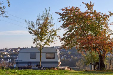 Charming Camper Amidst Autumn Landscape: A Cozy Retreat Surrounded by Trees, Stone Barbecue, and a Clear Blue Sky at a Summer Cottage in Germany. This picturesque scene captures a cozy camper nestled clipart