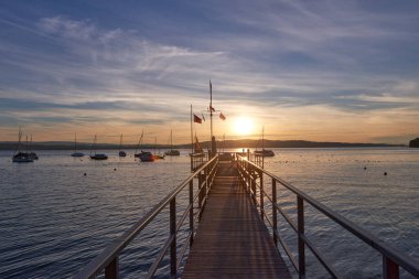 Sakin bir nehrin üzerinde sakin bir günbatımı, canlı turuncu ve pembe renkler gökyüzünde. Küçük bir yelkenli, rengarenk gökyüzünü yansıtan suya demir atmış..
