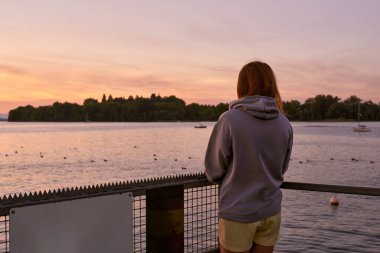 Bu çarpıcı görüntü, gölün kenarındaki iskelede duran güzel bir kızı, sarı şort ve gri bir kapüşonlu ile inanılmaz bir gün doğumuna bakarken yakalıyor. Sabah ilk ışıklarıyla