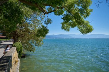 Lindau, Almanya 'daki Güzel Kız Gölü ve Dağ Manzarası: Constance Gölü' ndeki Yelkenliyle Yaz Huzuru. Bu büyüleyici görüntü gölün kıyısında duran güzel bir kızı gösteriyor.