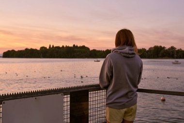 Kısa Şortlu Güzel Kadın ve Gri Kapüşonlu Şortlu Gri Başlıklı Şafak Gölü kıyısındaki bir rıhtımda, Şafakta Sersemletici Gündoğumu, Serene Gölü ve Gökyüzü Sahnesi, Sabah Atmosferi