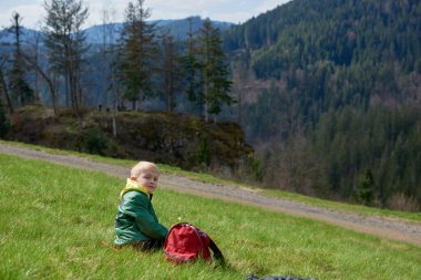 Eğlenceli genç yamaçta oturur. Çocuk dağlarda dinleniyor. Okul çocuğu çim tarlasında tepede oturuyor ve manzaranın tadını çıkarıyor. Doğada dinlenen ergen çocuk yürüyüşçüsünün arka görüntüsü. Etkin
