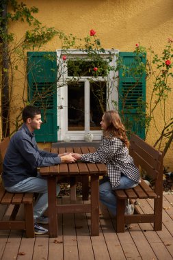 İki kişinin ahşap bir piknik masasında el ele tutuştuğu sakin bir bahçe sahnesi. Resimli dekorda yeşil panjurlu sarı bir bina, beyaz çerçeveli pencereler ve
