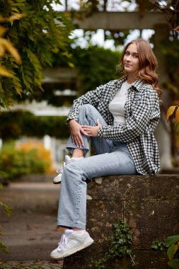 Şık bir genç kadın, canlı bir şehir parkındaki yıpranmış taş bir duvara tünemiş sonbahar zarafetini somutlaştırıyor. Çıtır çıtır siyah beyaz bir gömlek giyip klasik mavi kot pantolon giyip...