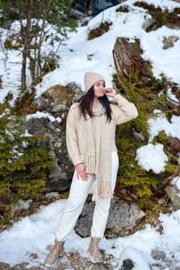 Yirmili yaşlarda şık krem rengi örgü ve rahat botlar giyen çekici bir kadın model, kış ormanı ortamında doğal olarak poz veriyor. Sıcak bere takan şık bir kışlık kıyafet.