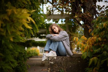 Güzel genç bir kadın, sakin bahçedeki beyaz ahşap bankta oturuyor, yemyeşil, siyah beyaz kareli ceket, mavi kot ve beyaz spor ayakkabılar da dahil olmak üzere günlük giysiler giyiyor.