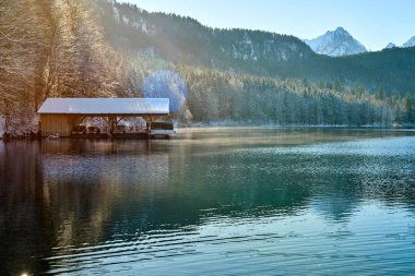Bu fotoğraf sakin bir göl kenarında sakin bir kış sahnesini çeker. Görüntü, kısmen karla kaplı bir çatı ile kaplı, suya uzanan ahşap bir rıhtıma sahip. Rıhtım ahşap sütunlarla destekleniyor ve demirlemiş birkaç küçük tekne var.