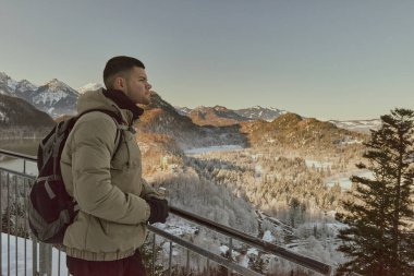 Balkonda güven içinde duran, muhteşem buzdan dağlara ve arka planda yemyeşil bir ormana bakan bir adamın çarpıcı görüntüsü. Fotoğraf, portre şeklinde çekiliyor, karla kaplı, nefes kesen kış manzarasını yakalıyor.