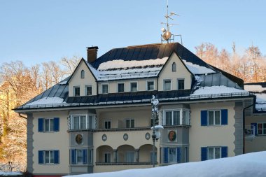 Bu fotoğraf, koyu mavi, karla kaplı çatısı olan çok katlı, büyüleyici bir konut binasının olduğu huzurlu bir kış sahnesini çekiyor. Yumuşak, açık bej renkte boyanmış binanın mavi panjurlu ve küçük balkonlu birkaç büyük penceresi var.