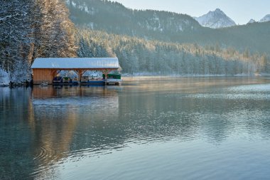Bu yüksek çözünürlüklü fotoğraf sakin bir göl kenarında sakin bir kış manzarası yakalar. Karla kaplı ahşap bir rıhtım, altında demirlemiş tekneleri destekleyen sütunları cam sulara kadar uzanıyor. Ön planda iki ördek var. Birinin parlak mavi kafası var.