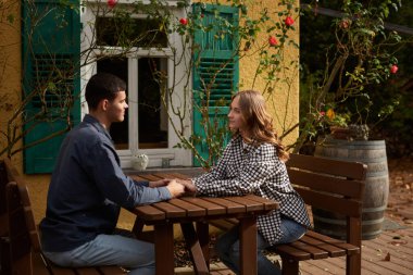 İki kişinin ahşap bir piknik masasında el ele tutuştuğu sakin bir bahçe sahnesi. Resimli dekorda yeşil panjurlu sarı bir bina, beyaz çerçeveli pencereler ve