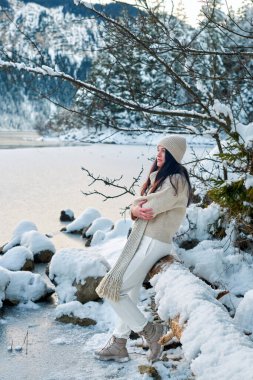 Uzun siyah saçlı, bej örgü şapka, krem rengi örgü eşarp, krem rengi kazak, beyaz tayt ve ten rengi ayakkabı giyen bir kadının yer aldığı sakin bir kış sahnesi. Karın üzerinde duruyor.