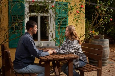 İki kişinin ahşap bir piknik masasında el ele tutuştuğu sakin bir bahçe sahnesi. Resimli dekorda yeşil panjurlu sarı bir bina, beyaz çerçeveli pencereler ve