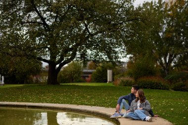 Genç bir çift, güzel bir sonbahar parkında, canlı bir sonbahar yeşillikleriyle çevrili bir göletin kenarında yan yana otururlar. Tarz sahibi, kareli gömlek ve kot giyen kadın nazikçe eğildi.