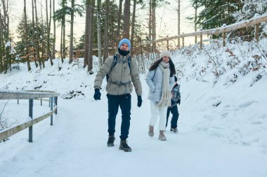 Bu yüksek çözünürlüklü fotoğraf, kışın karlı bir ormanda yürüyen bir ailenin yürek ısıtan bir görüntüsünü çeker. Bir erkek, bir kadın ve bir çocuktan oluşan aile dinginliğin tadını çıkarıyor.