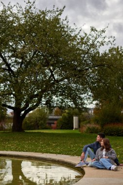 Bu pitoresk sahne, sakin bir park veya bahçedeki yansıtıcı bir göletin kenarında çimlerin üzerinde oturan iki kişiyi yakalar. Çift birbirine yaslanıyor ve bir anlığına eğleniyor.