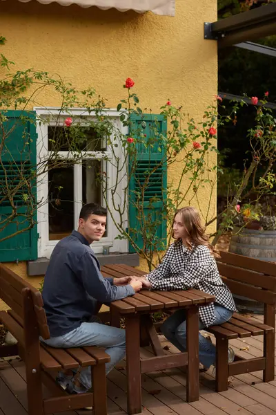 Bir çiftin geleneksel yeşil panjurlarla süslenmiş klasik sarı bir binanın yanına kurulmuş büyüleyici bir bahçe piknik masasında el ele tutuşması gibi hassas bir an yaşanıyor. Sahne yemyeşil bir gül tarafından çerçevelenmiş.