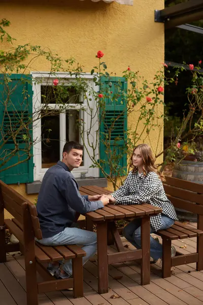 İki kişinin ahşap bir piknik masasında el ele tutuştuğu sakin bir bahçe sahnesi. Resimli dekorda yeşil panjurlu sarı bir bina, beyaz çerçeveli pencereler ve