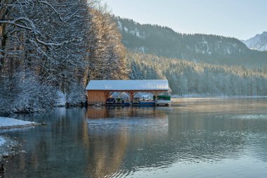 Bu yüksek çözünürlüklü fotoğraf sakin bir göl kenarında sakin bir kış manzarası yakalar. Karla kaplı ahşap bir rıhtım, altında demirlemiş tekneleri destekleyen sütunları cam sulara kadar uzanıyor. Ön planda iki ördek var. Birinin parlak mavi kafası var.