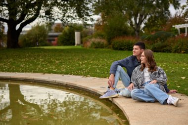 Bu pitoresk sahne, sakin bir park veya bahçedeki yansıtıcı bir göletin kenarında çimlerin üzerinde oturan iki kişiyi yakalar. Çift birbirine yaslanıyor, sonbaharın canlı renkleriyle çevrili bir rahatlama anının tadını çıkarıyor.