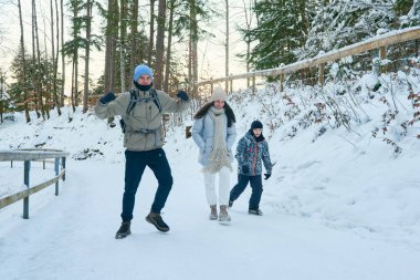 Bu yüksek çözünürlüklü fotoğraf, kışın karlı bir ormanda yürüyen bir ailenin yürek ısıtan bir görüntüsünü çeker. Bir erkek, bir kadın ve bir çocuktan oluşan aile, buzlu yolda yürürken huzurlu ve pitoresk manzaranın tadını çıkarıyor.