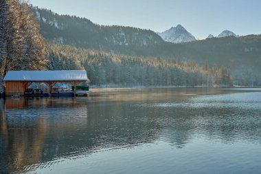 Bu yüksek çözünürlüklü fotoğraf dingin bir göl kıyısında nefes kesici bir kış gündoğumunu yakalıyor. Odak noktası kırsal ahşap bir kayıkhane. Kısmen cam sulara batırılmış. Çevredeki manzarayı mükemmel yansıtıyor. Kar battaniyeleri